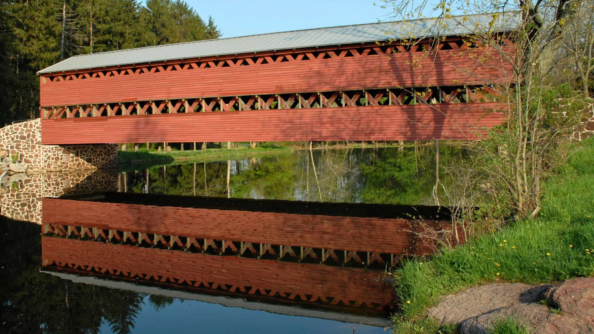 A long red wooden covered bridge spans across calm water, creating a perfect mirror reflection, with stone abutments and green trees surrounding it.