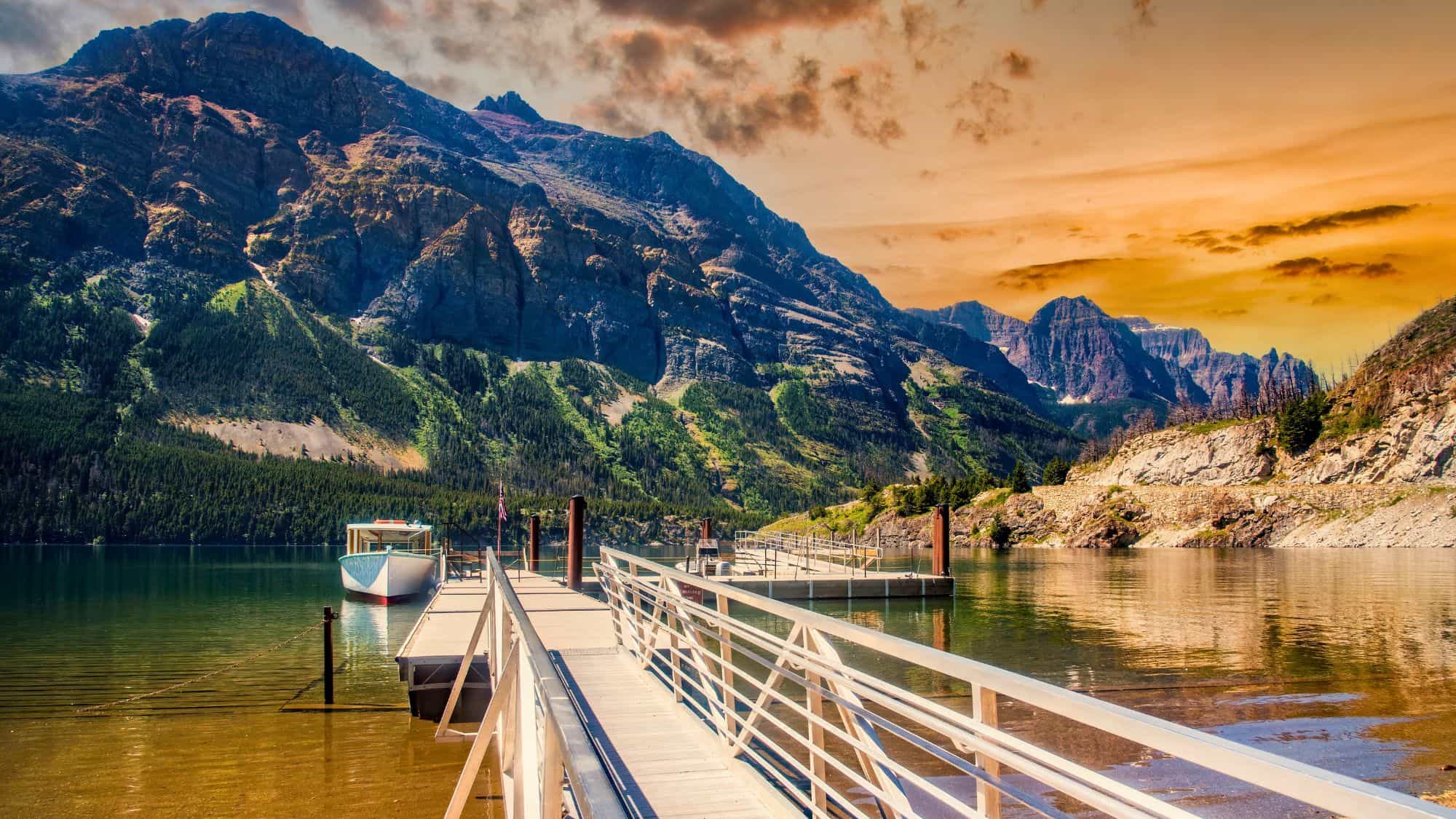 A scenic dock stretches into the clear waters of Saint Mary Lake, with towering mountains and a dramatic sunset sky glowing orange in the background.