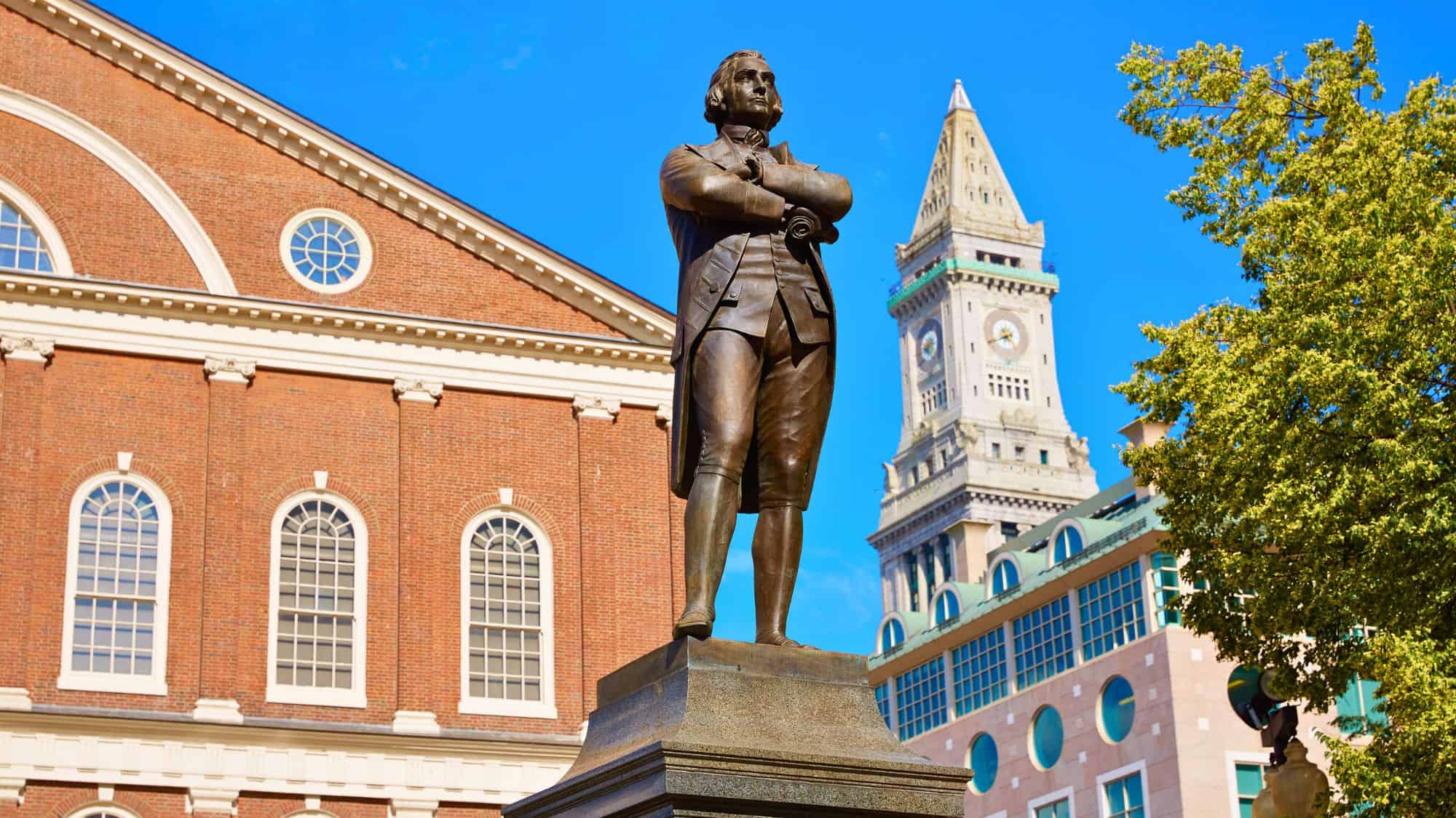 A bronze statue of Samuel Adams stands proudly with arms crossed in front of Boston’s historic Faneuil Hall, with the Custom House Tower visible in the background under a clear blue sky.