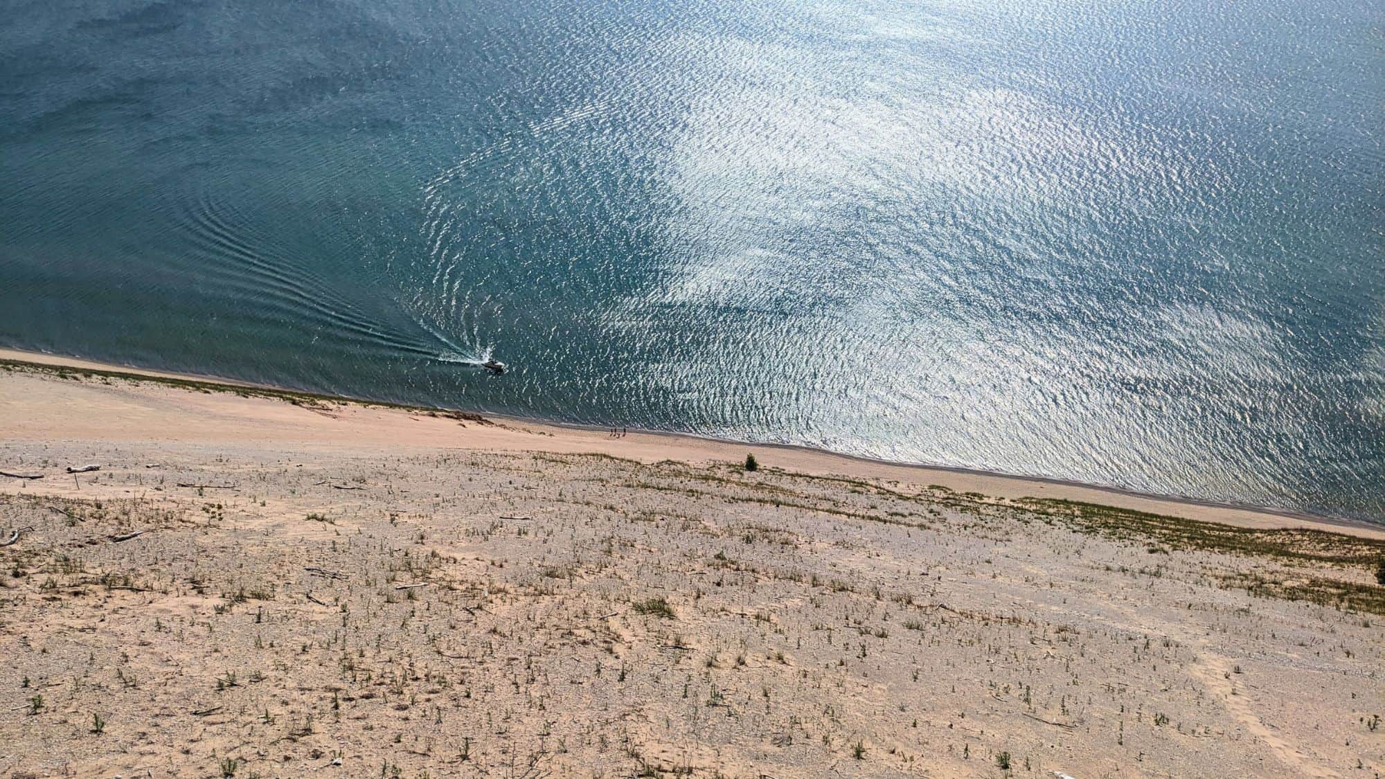 A wide view from the top of Sleeping Bear Dunes shows a steep sandy slope leading down to the shimmering blue waters of Lake Michigan, with a small boat cutting across the water below.