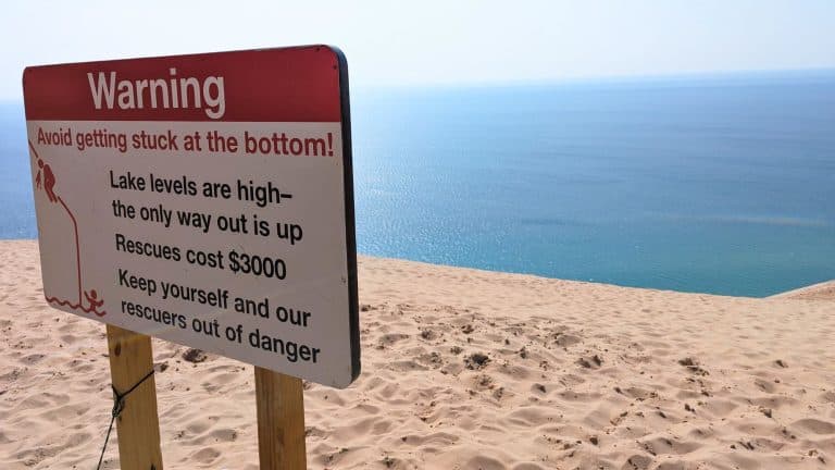 A red-and-white warning sign at the top of Sleeping Bear Dunes cautions hikers about high lake levels and the cost of rescue, with expansive blue water and sand stretching out behind it.