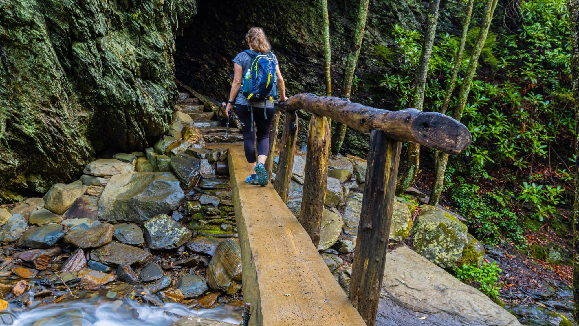 A woman with a backpack and hiking poles walks across a narrow wooden footbridge surrounded by mossy rocks and dense green foliage in the Smoky Mountains.