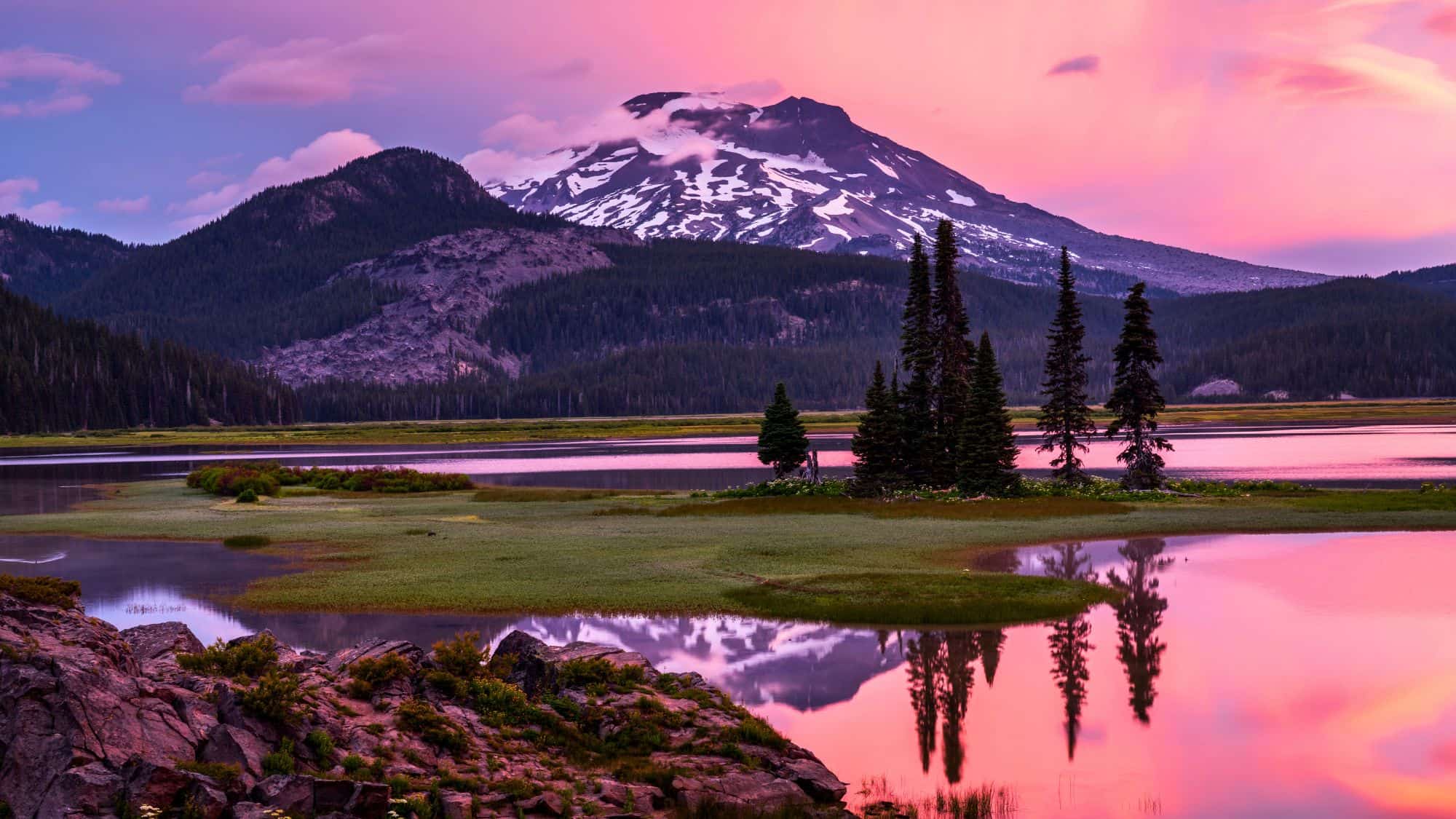 A breathtaking view of Sparks Lake at sunset, with vibrant pink and purple skies framing a snow-capped mountain and mirrored reflections in the still water.