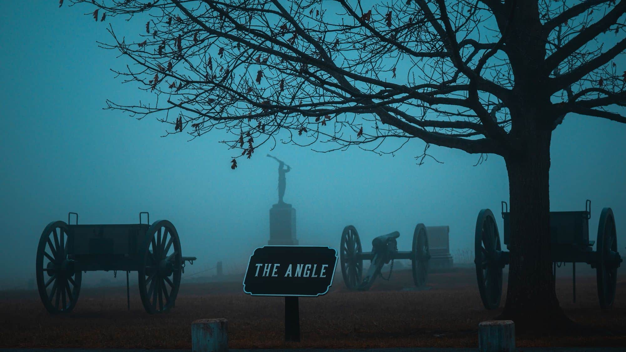 A foggy battlefield scene at Gettysburg National Military Park featuring Civil War-era cannons, a bare tree, and a sign labeled “The Angle,” with a lone statue in the background.