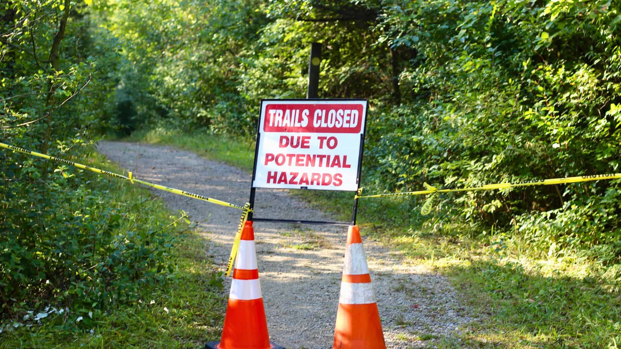 A hiking path blocked off with yellow caution tape and orange cones, featuring a red and white sign that reads "TRAILS CLOSED DUE TO POTENTIAL HAZARDS."