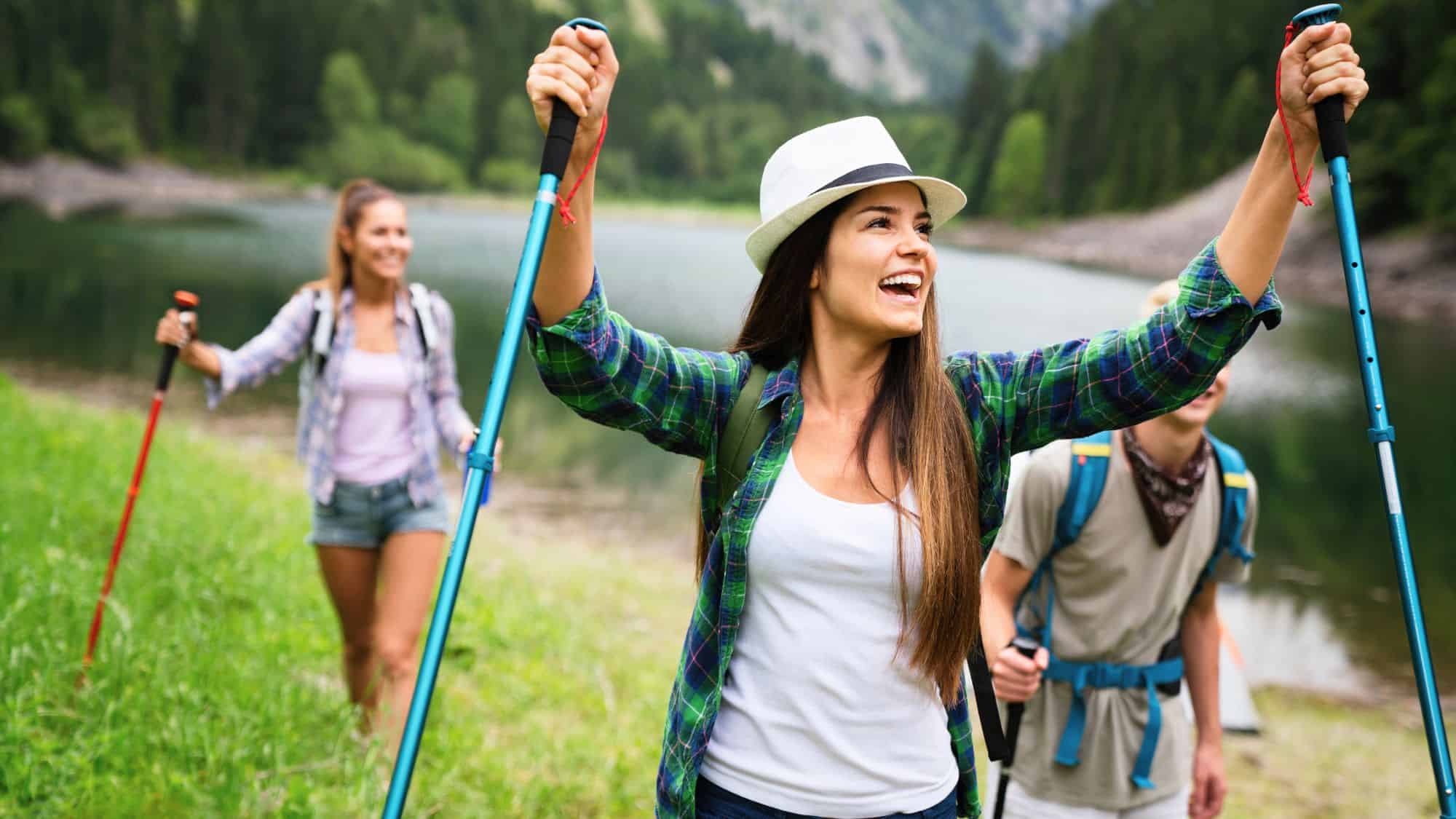 A smiling woman in a plaid shirt raises her trekking poles in celebration while hiking with friends near a mountain lake, surrounded by green trees and hills.