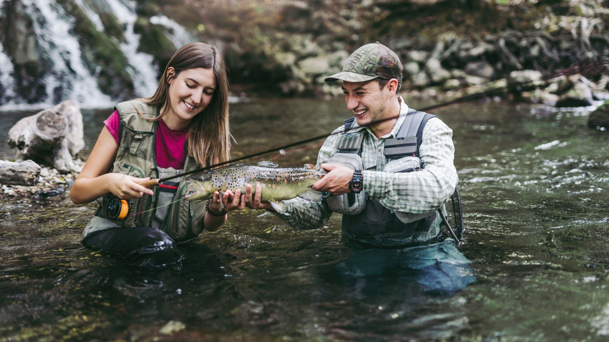 A smiling young couple standing knee-deep in a clear stream proudly hold a large speckled trout between them, with a waterfall and rocky terrain in the background.