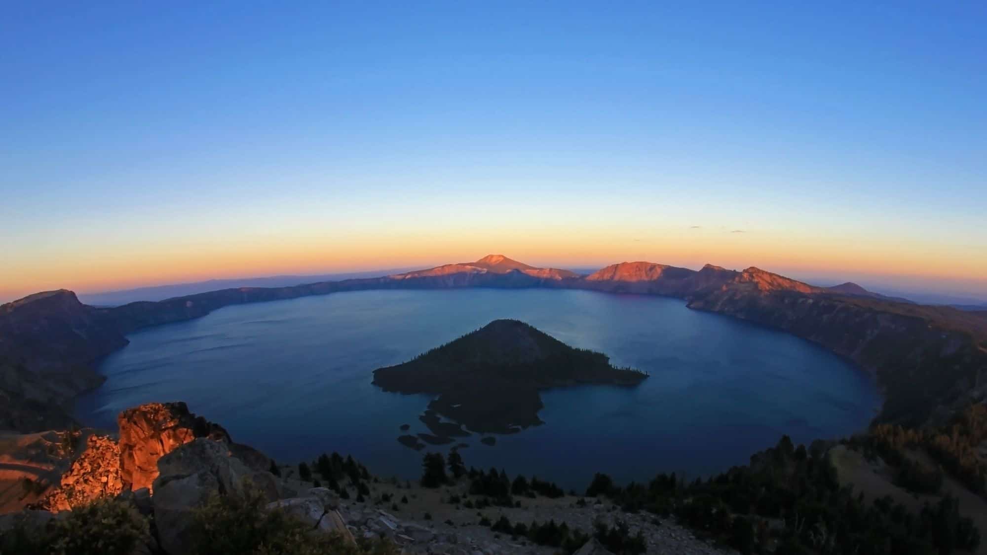 Sunrise over Crater Lake viewed from Watchman’s Peak, with the lake’s central Wizard Island silhouetted against glowing orange and pink mountain ridges.