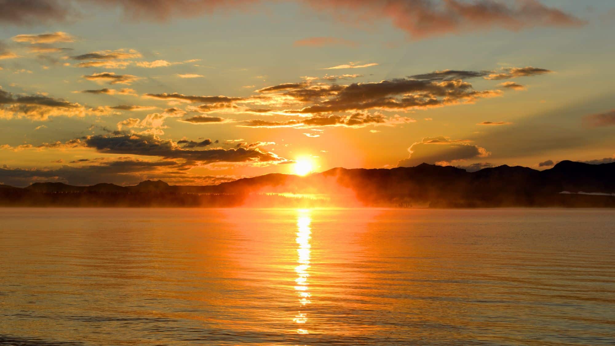 A fiery orange sunrise lights up Yellowstone Lake, with mist rising off the water and silhouetted mountains in the distance.