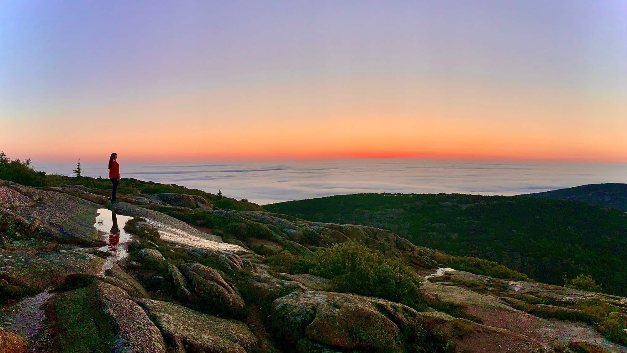 A lone hiker stands on the rocky summit of Cadillac Mountain in Acadia National Park, gazing at a serene sunrise over a sea of low-lying clouds and forested hills.