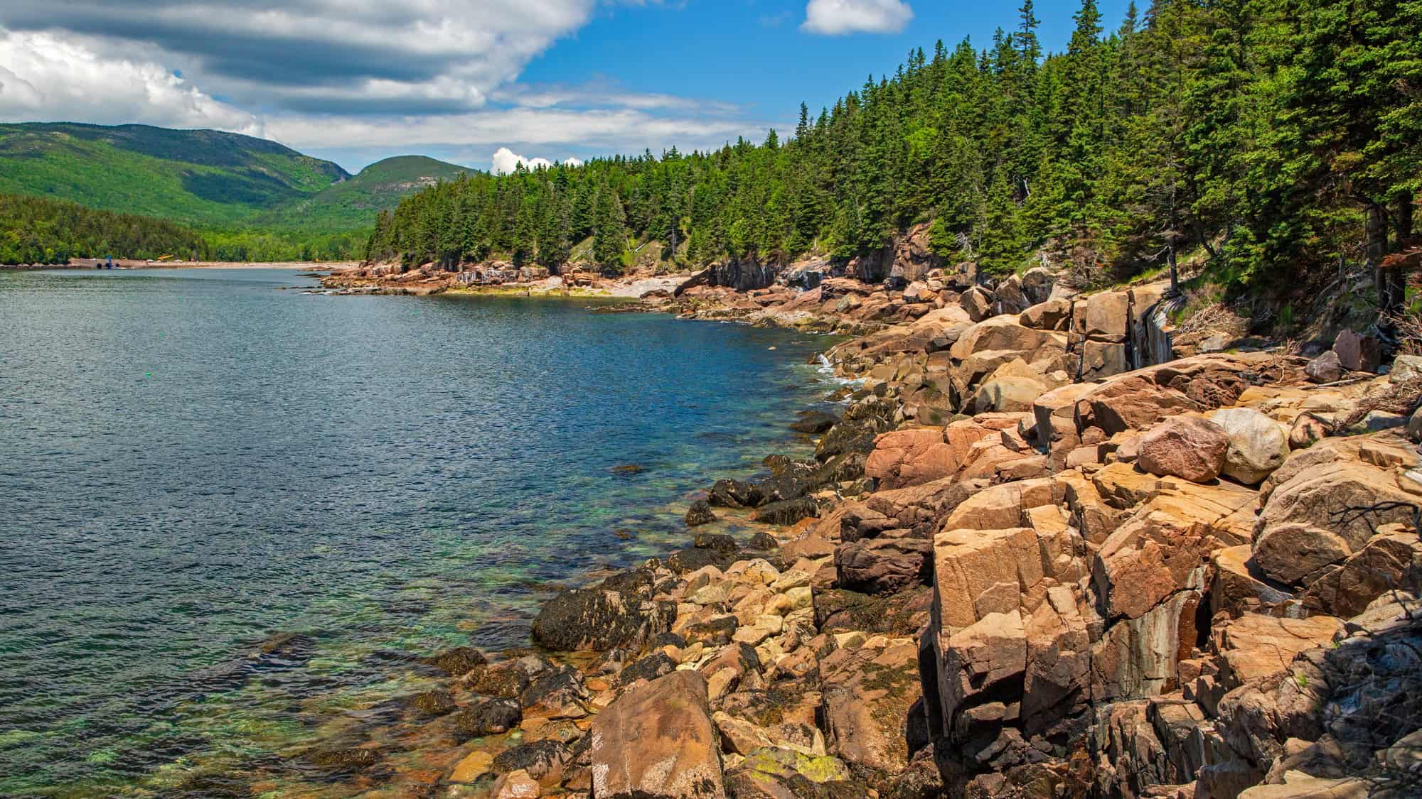 The rugged coastline of Acadia National Park features rocky outcrops and forested slopes beside crystal-clear waters, with green mountains rising in the background.