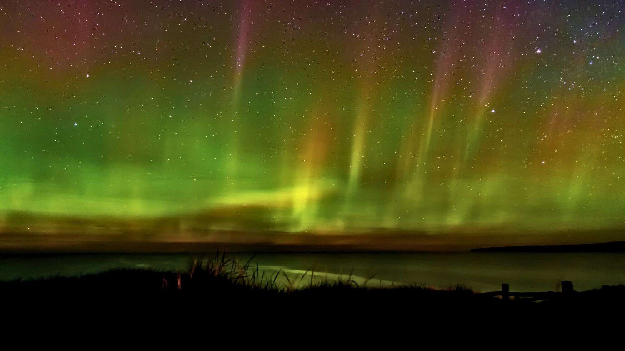 Dancing ribbons of green and faint red aurora borealis light up the night sky over Lake Superior, with stars sparkling across the scene and a quiet silhouette of the shoreline below.
