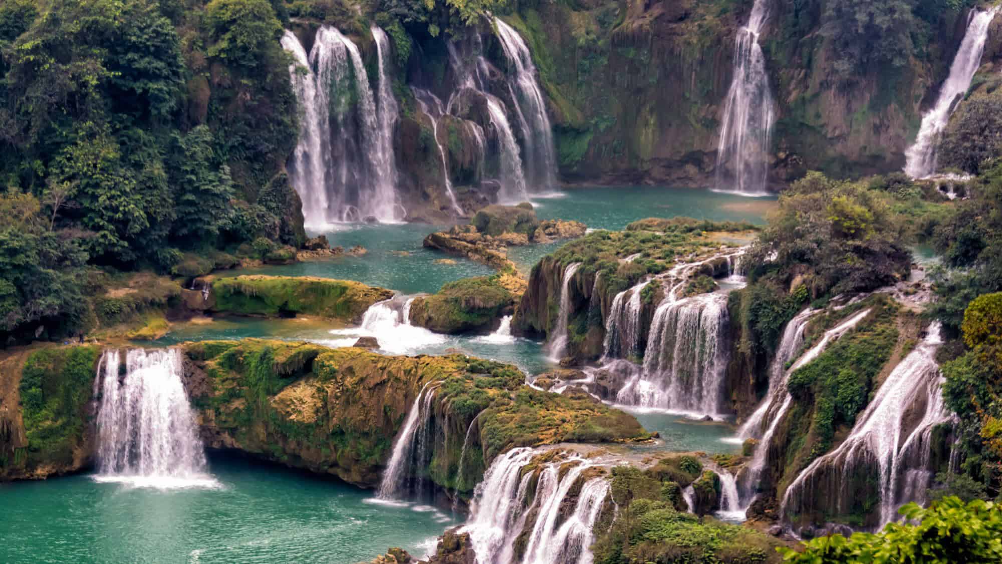 A complex network of waterfalls cascades over tiered limestone rocks into emerald green pools, surrounded by dense tropical forest.
