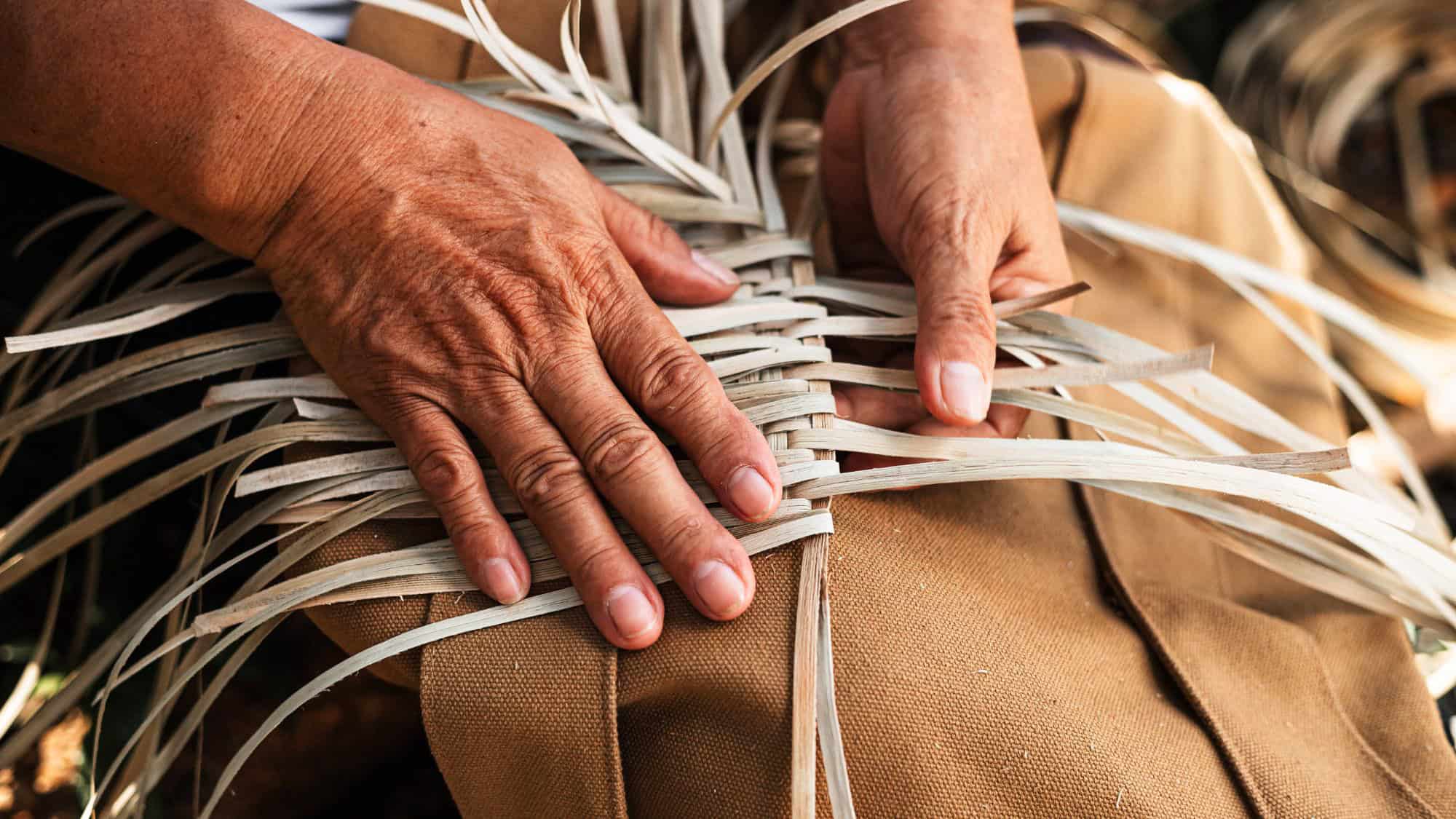 Close-up of experienced hands weaving thin, pale reeds into a basket, showing the intricate craftsmanship and texture of the materials against a tan fabric background.