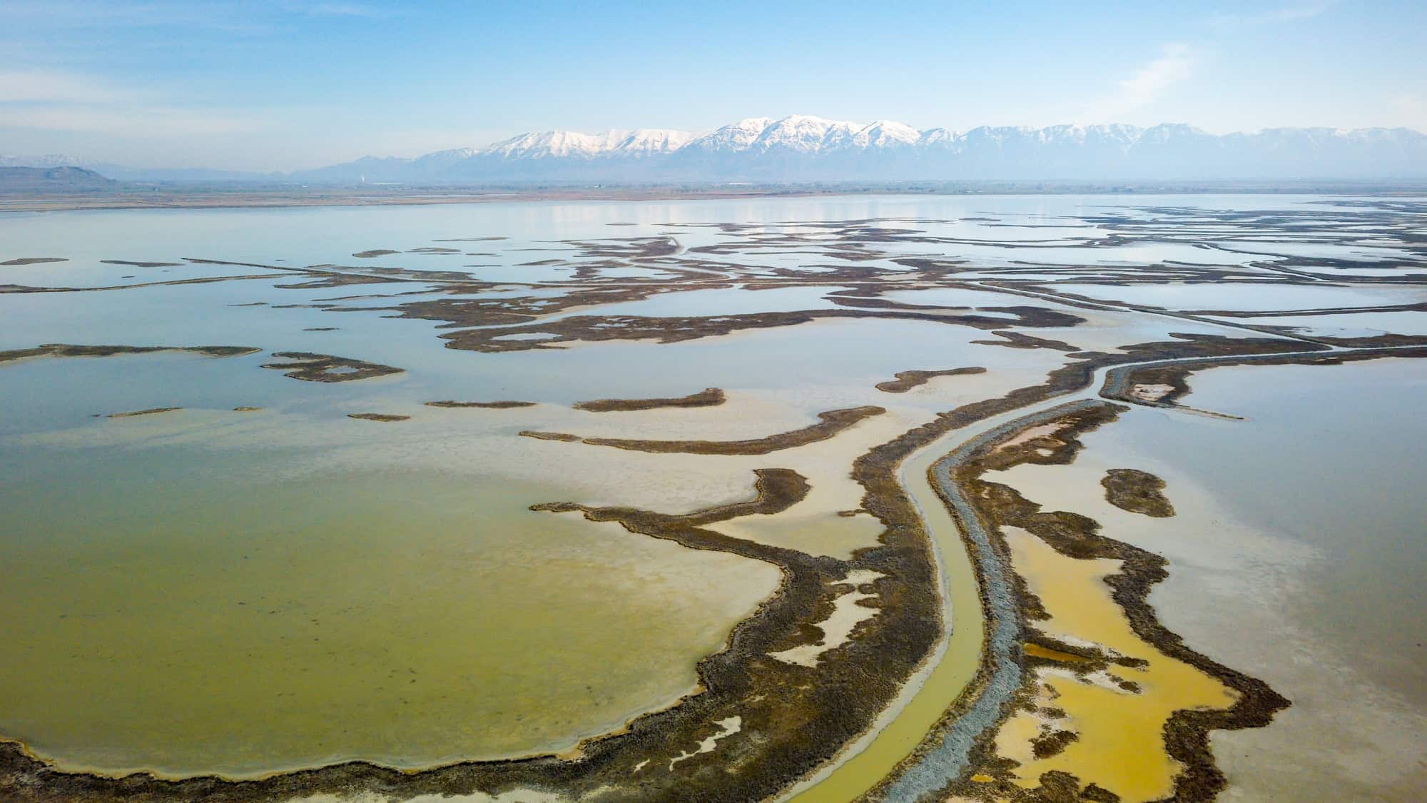 A sweeping aerial view of Bear River Migratory Bird Refuge shows winding wetland channels and tan mudflats stretching into the distance, with a snowcapped mountain range rising under a clear blue sky.
