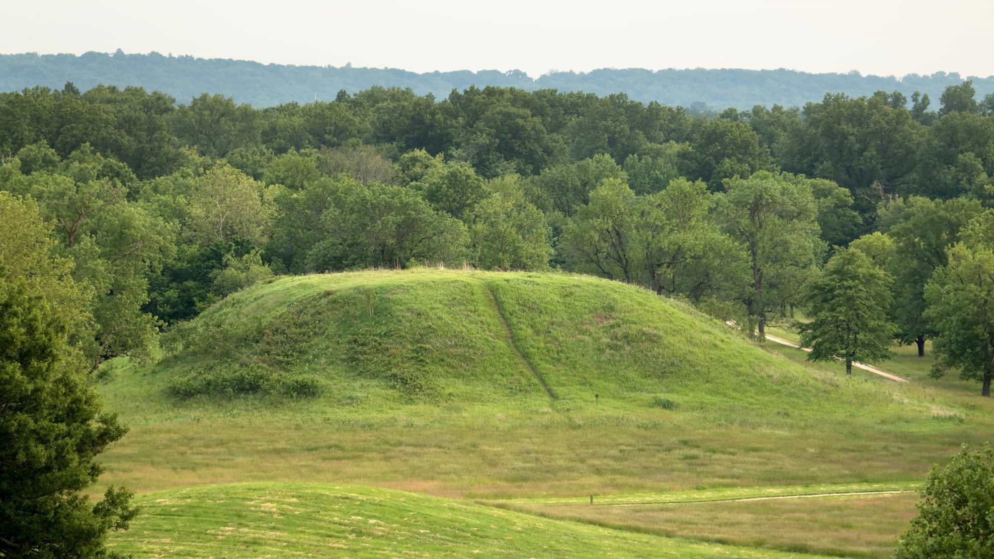 A large, grassy mound rises from a field in southern Illinois, surrounded by forest—part of a pre-Columbian Native American city and UNESCO World Heritage Site.