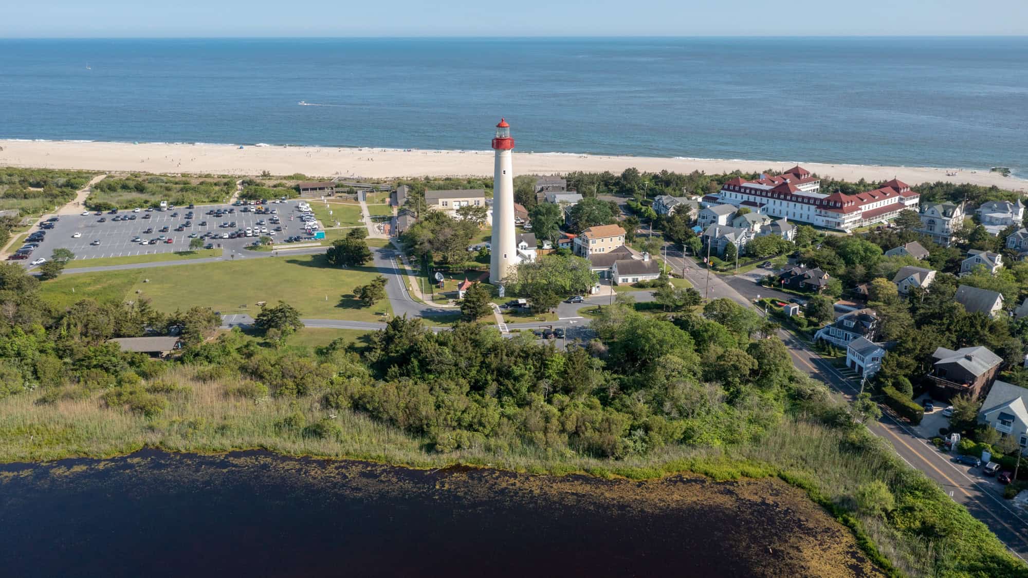 An overhead shot of Cape May, New Jersey, centers on the iconic red-topped lighthouse near the beach, surrounded by a quaint seaside community and parking lot