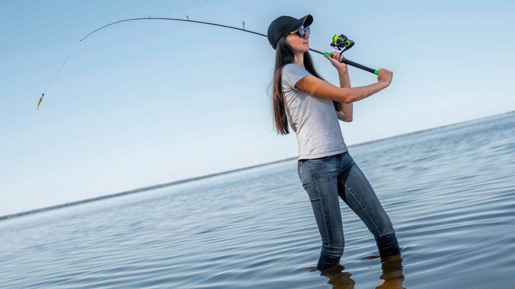 A woman wearing sunglasses and a baseball cap stands knee-deep in water, skillfully casting a fishing line under a bright blue sky.