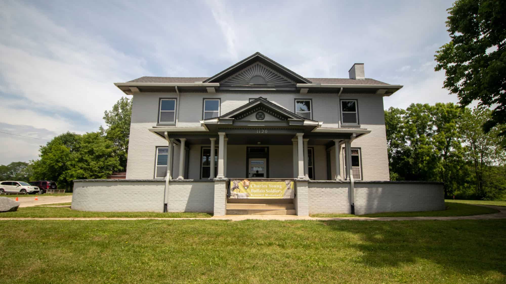 A two-story gray brick house with white trim and a porch supported by columns. A banner across the front reads “Charles Young Buffalo Soldiers National Monument.”