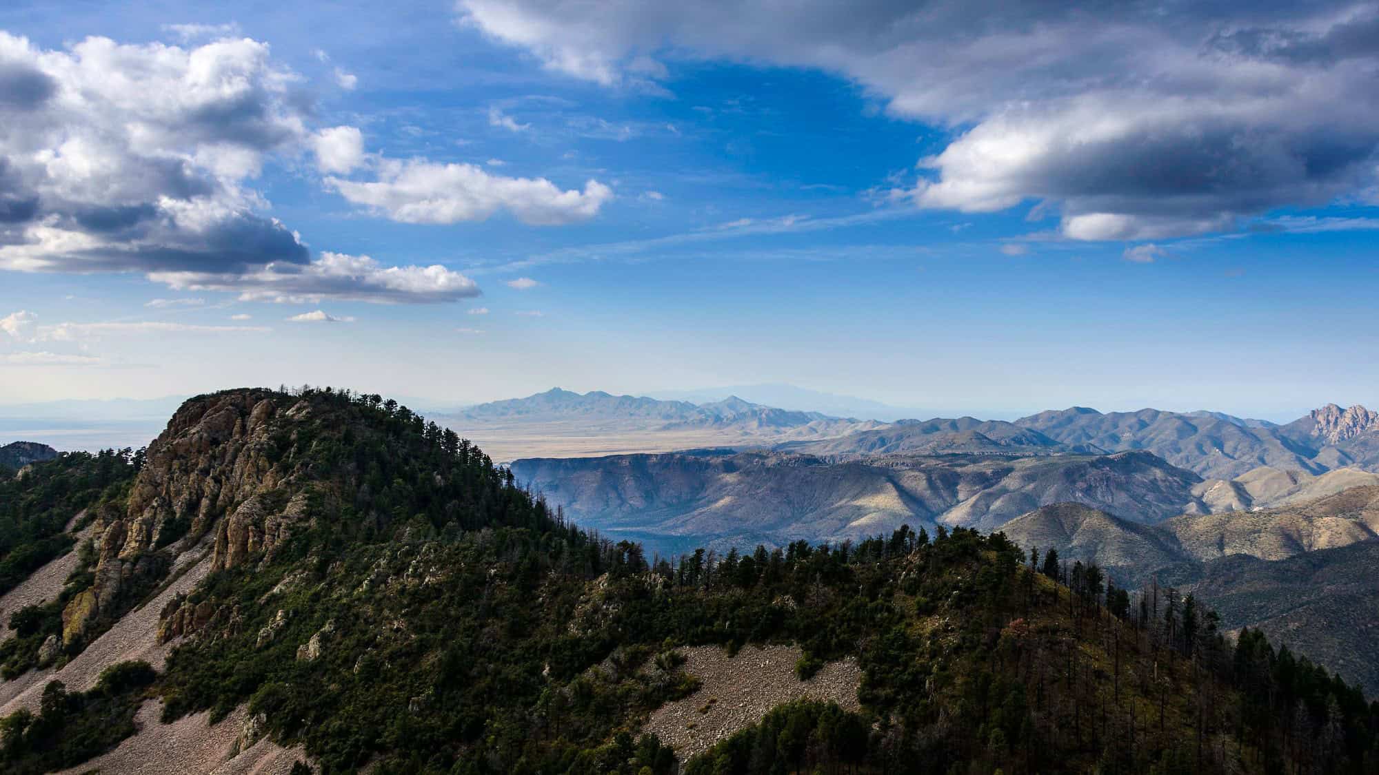 Rolling ridges and rocky peaks of the Chiricahua Mountains stretch toward the horizon under a partly cloudy sky, with deep valleys creating dramatic shadows.