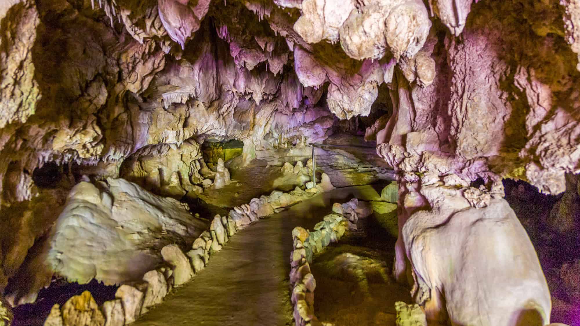 A winding path inside Crystal Cave lined with rocks and lit by purples and yellows, weaving through surreal stalagmites and hanging rock formations.