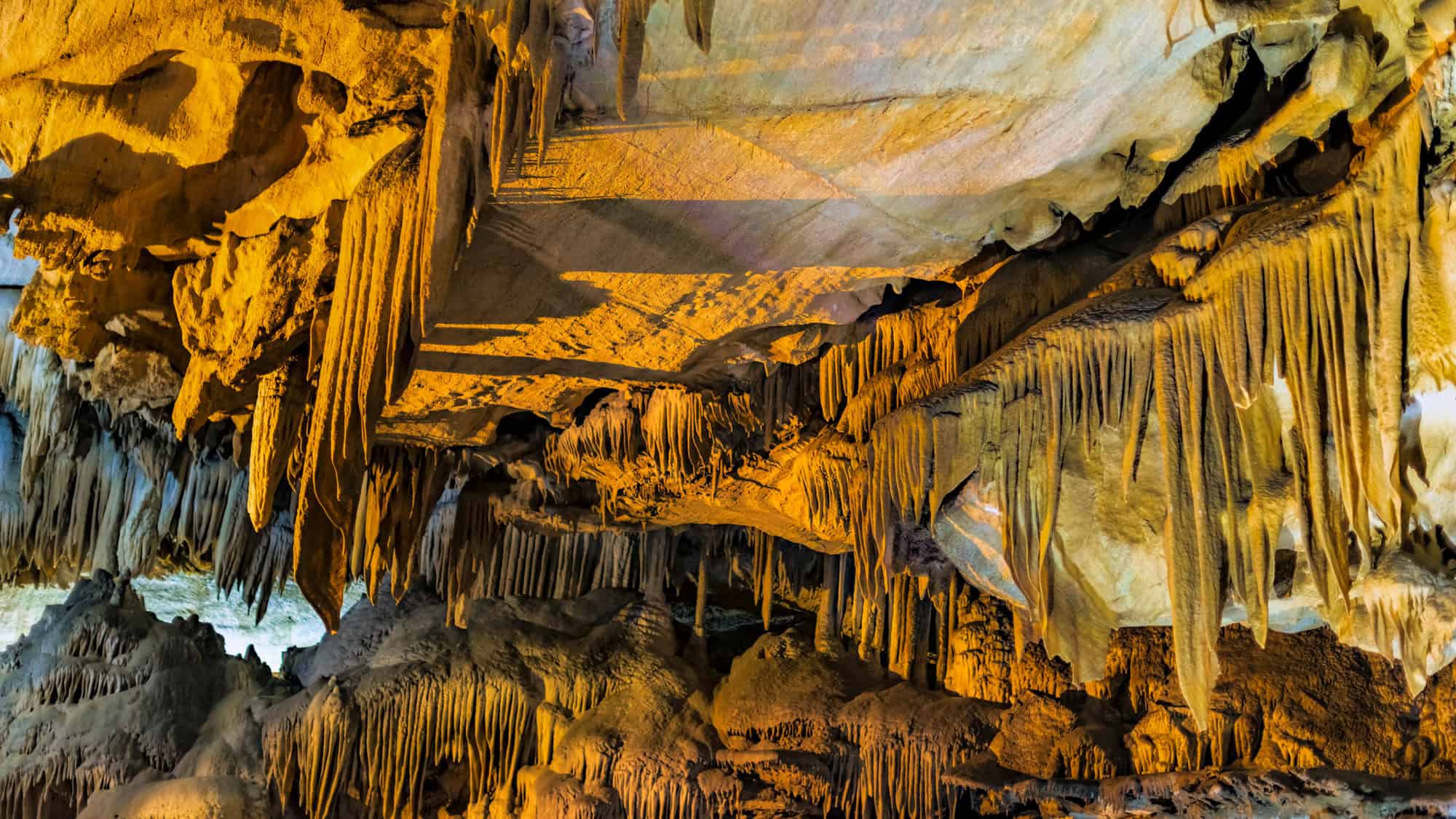 Sharp, jagged stalactites hang dramatically from a richly colored cave ceiling, illuminated by warm golden light that enhances the intricate mineral textures and shadows.