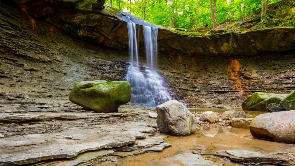 A delicate waterfall spills over a rock ledge into a shallow pool, framed by mossy boulders and green forest. This is Blue Hen Falls in Cuyahoga Valley National Park