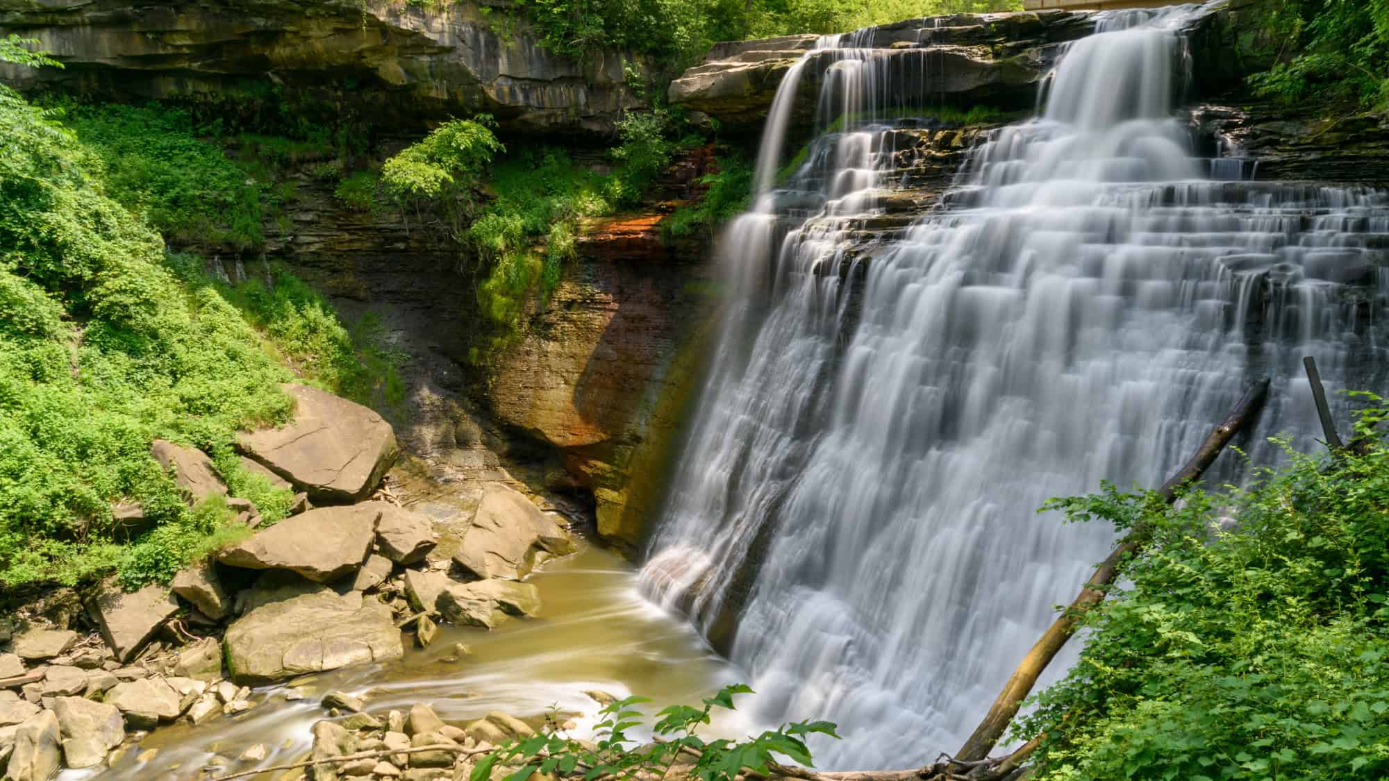 A powerful waterfall cascades down layered rock cliffs into a rocky stream below, surrounded by lush greenery in Cuyahoga Valley National Park.