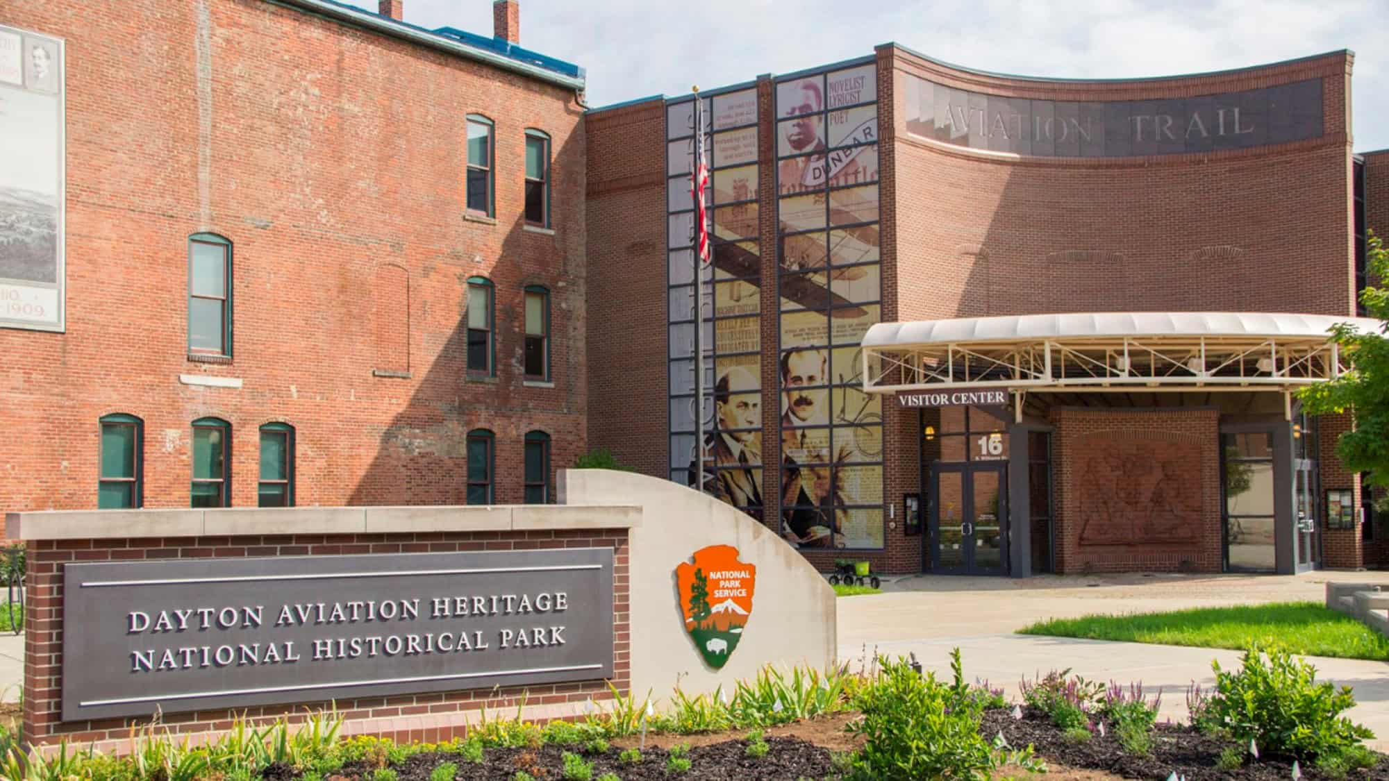 A brick building complex featuring a visitor center with “Aviation Trail” signage and murals of the Wright brothers. A sign out front reads “Dayton Aviation Heritage National Historical Park.”