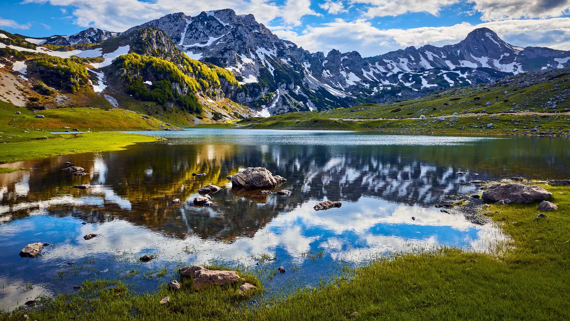 A serene alpine lake reflects rocky, snow-dusted peaks and patches of lush greenery under a vibrant blue sky in Montenegro’s Durmitor National Park.
