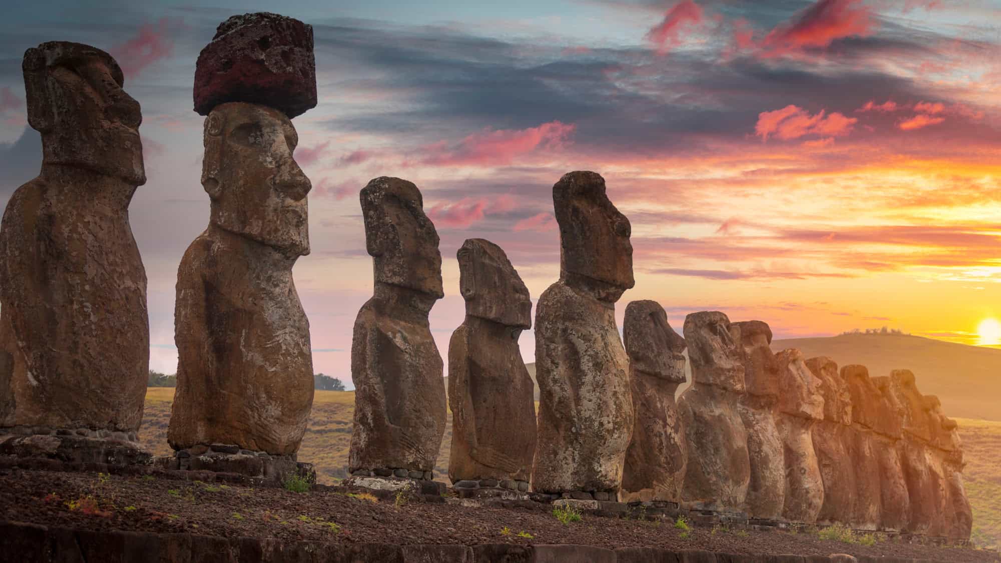 Row of towering Moai statues on Easter Island silhouetted against a colorful sunset sky with soft pink and orange hues.