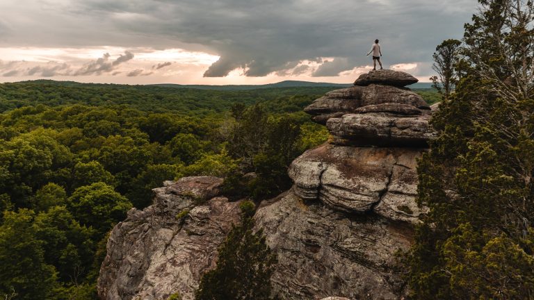 A lone hiker stands atop a massive sandstone rock formation, looking over a sweeping forested landscape under a dramatic, cloud-filled sky at sunset.