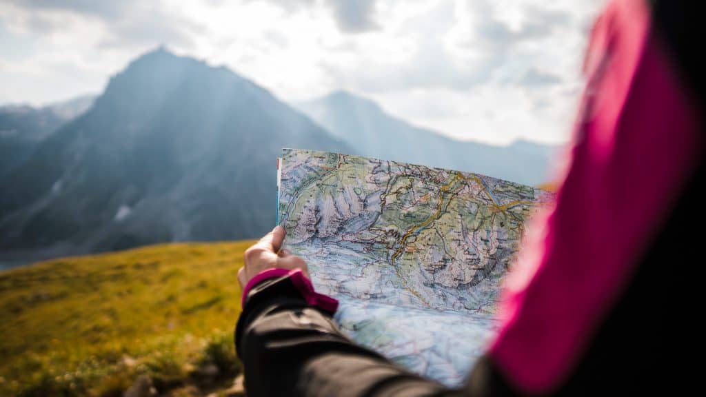 A person in a pink and black jacket holds a detailed topographic map with mountainous terrain and a misty, sunlit mountain range in the background.