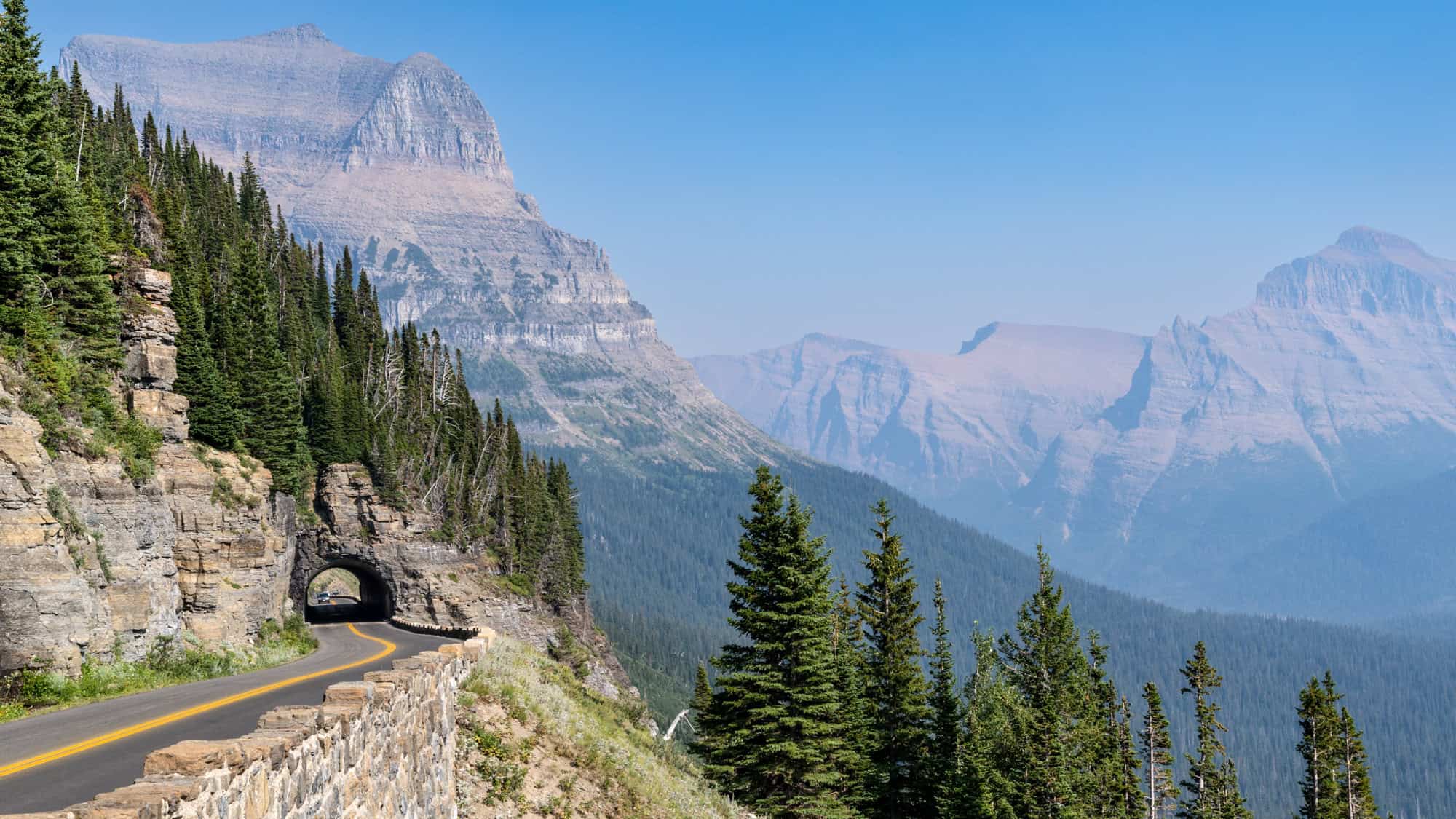 A scenic section of Glacier National Park's Going-to-the-Sun Road curves along a cliffside, passing through a rock tunnel with layered mountain peaks in the distance.