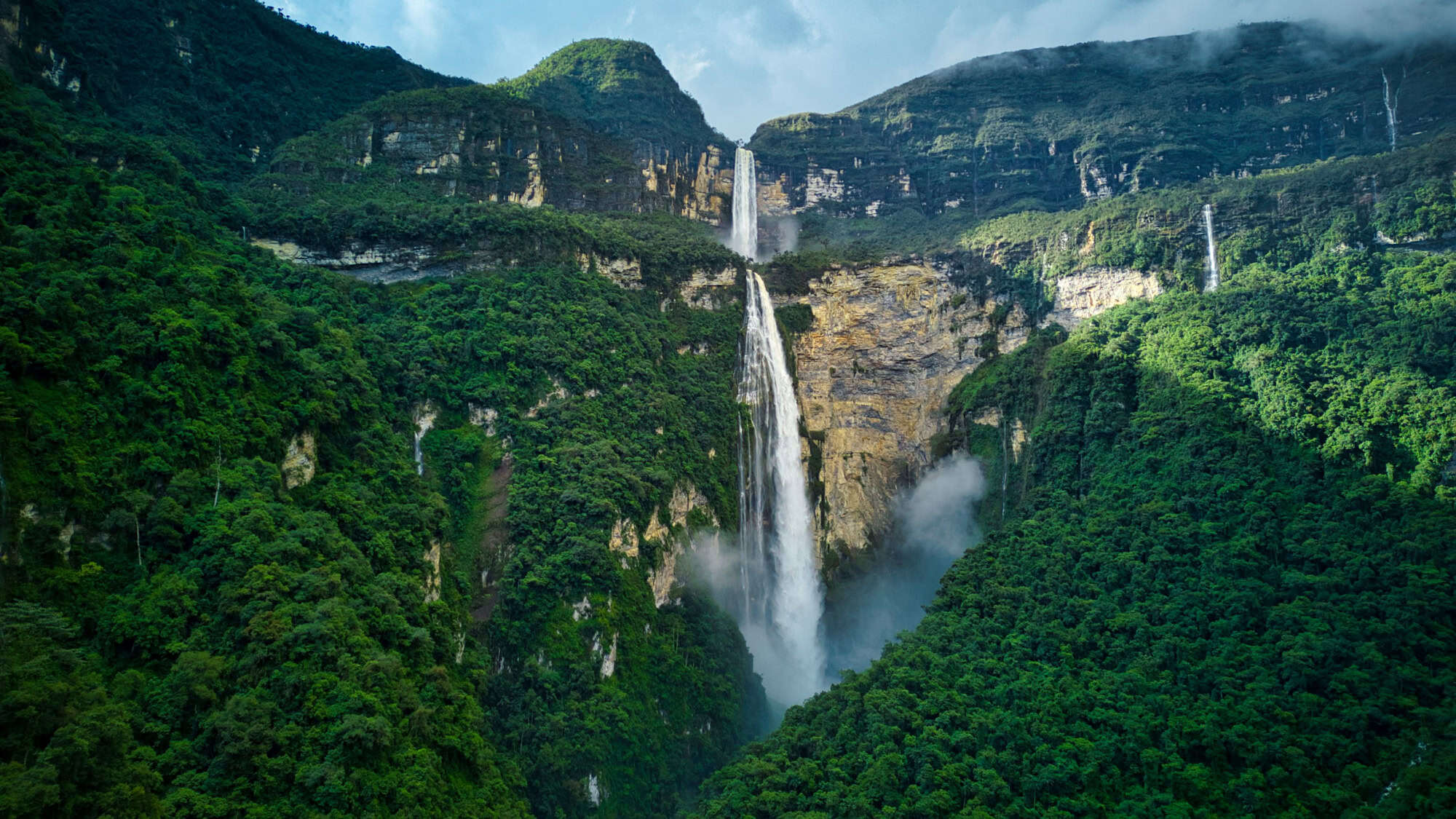 Gocta Falls in Peru pours dramatically down a massive cliff in two main drops, surrounded by thick green jungle and cloud-kissed mountain peaks. Mist rises from the waterfall’s base, adding to its powerful presence.
