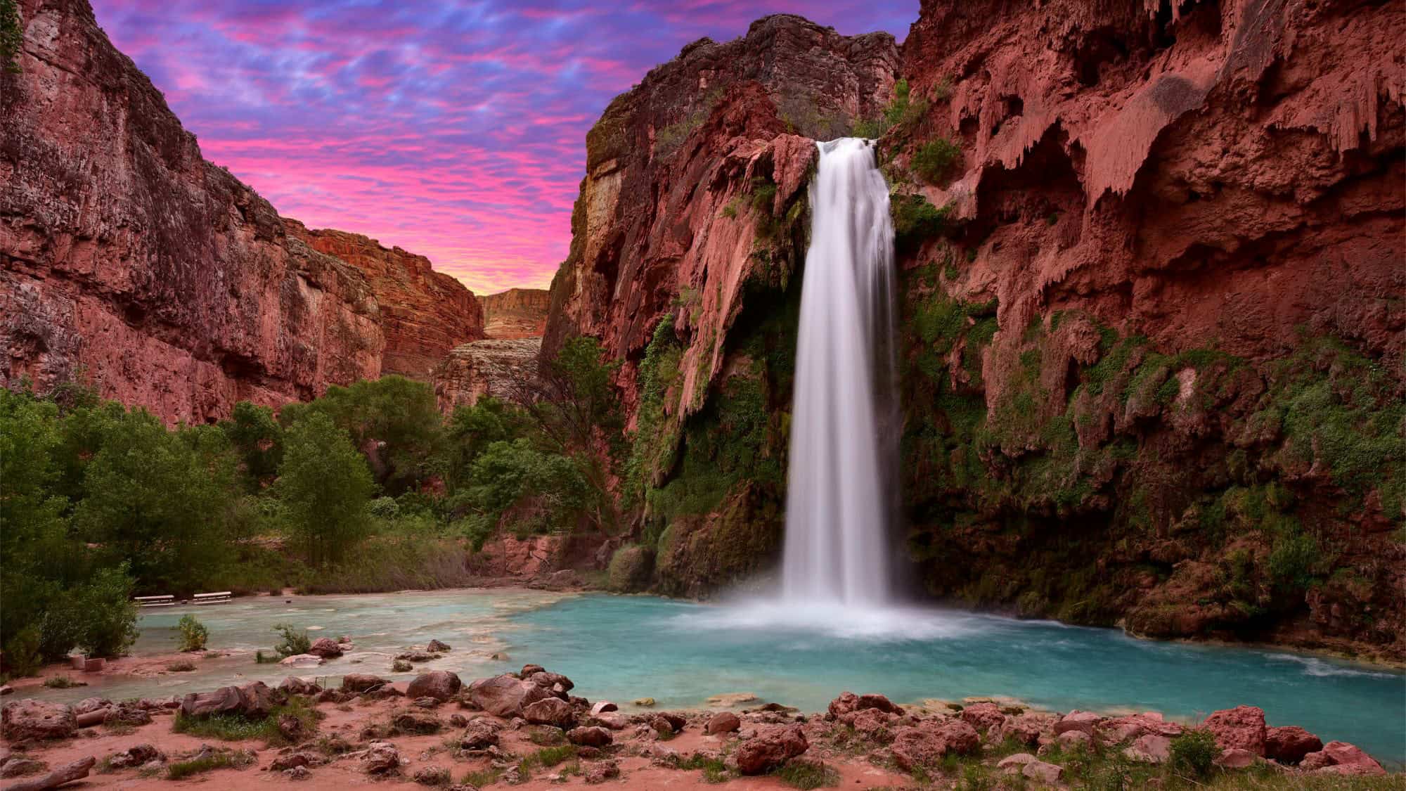 A powerful waterfall plunges into a turquoise pool, surrounded by red rock canyon walls and a vibrant pink and purple sunset sky.