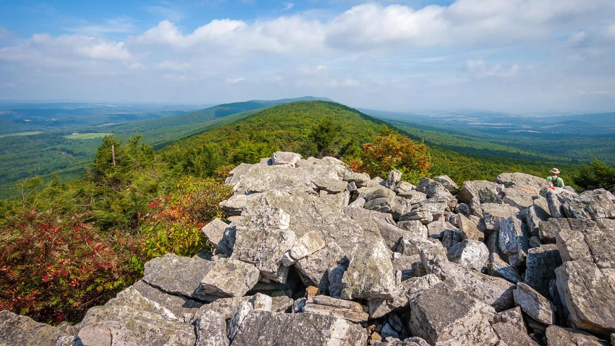From the summit of Hawk Mountain Sanctuary, a rocky outcrop overlooks a sweeping view of tree-covered ridges under a blue sky dotted with soft clouds.