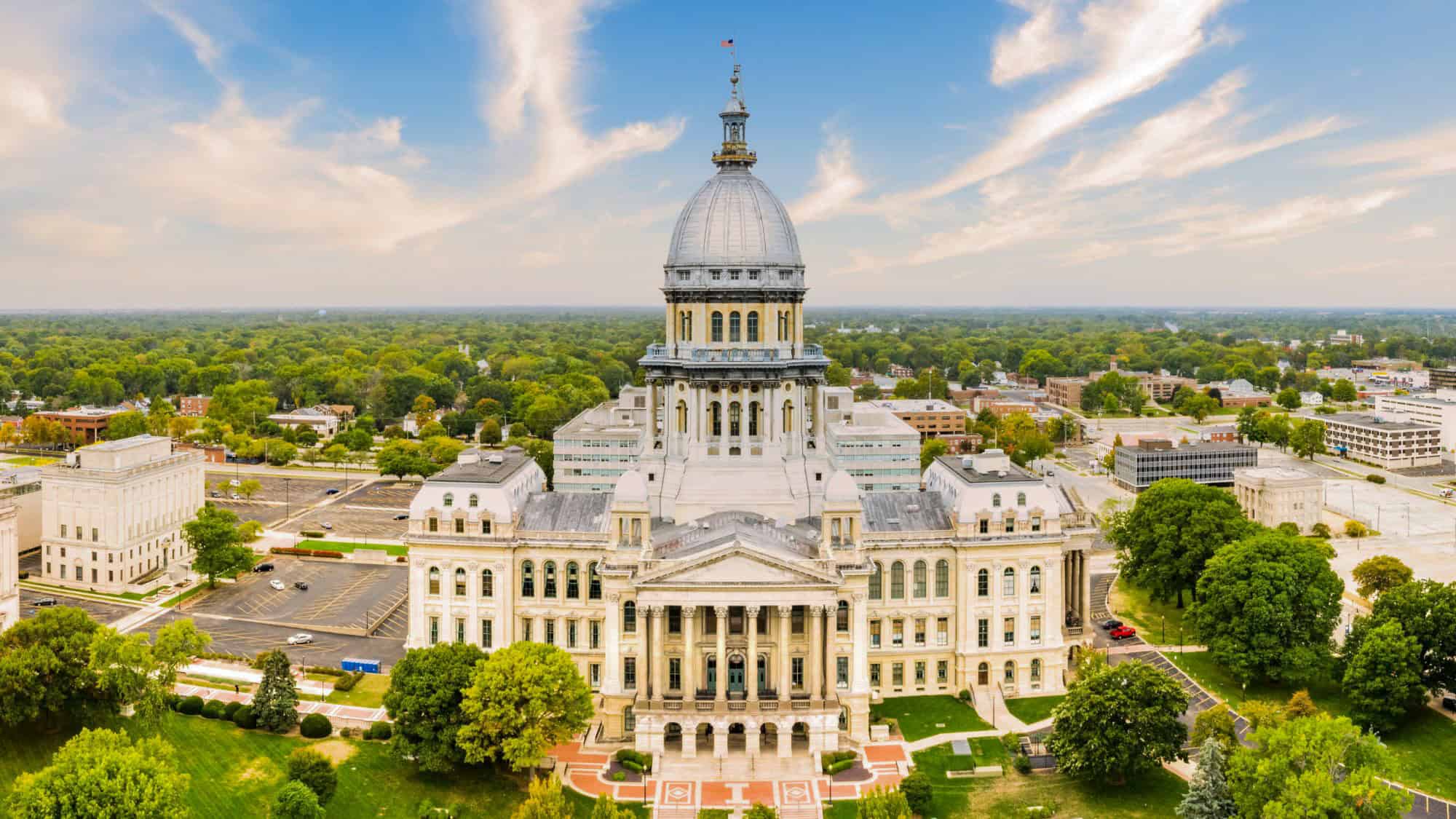 Aerial view of the grand Illinois State Capitol in Springfield, showcasing its majestic dome, ornate white stone façade, and surrounding cityscape beneath a partly cloudy sky.