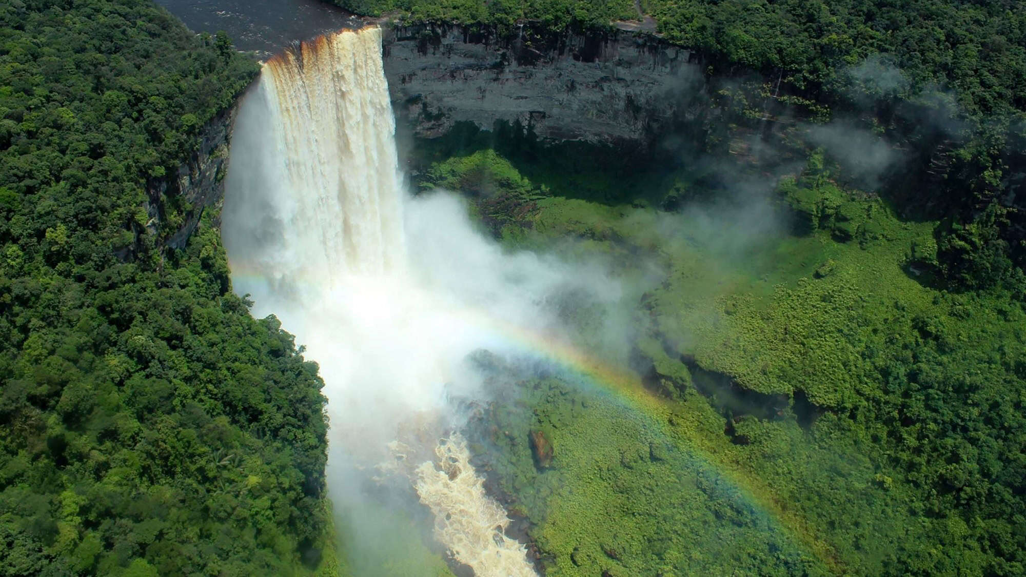 Aerial view of Kaieteur Falls dropping dramatically into a jungle gorge, with a rainbow forming in the mist at the base of the falls.