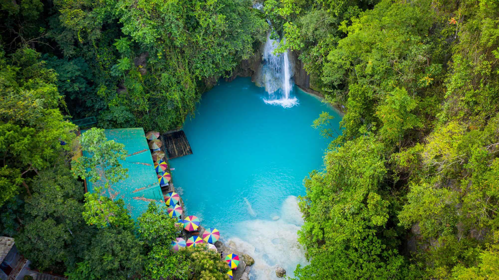 A vibrant aerial shot of Kawasan Falls in Cebu, Philippines, showing a small waterfall cascading into a bright turquoise pool, surrounded by dense tropical greenery and colorful umbrellas lined along the edge.