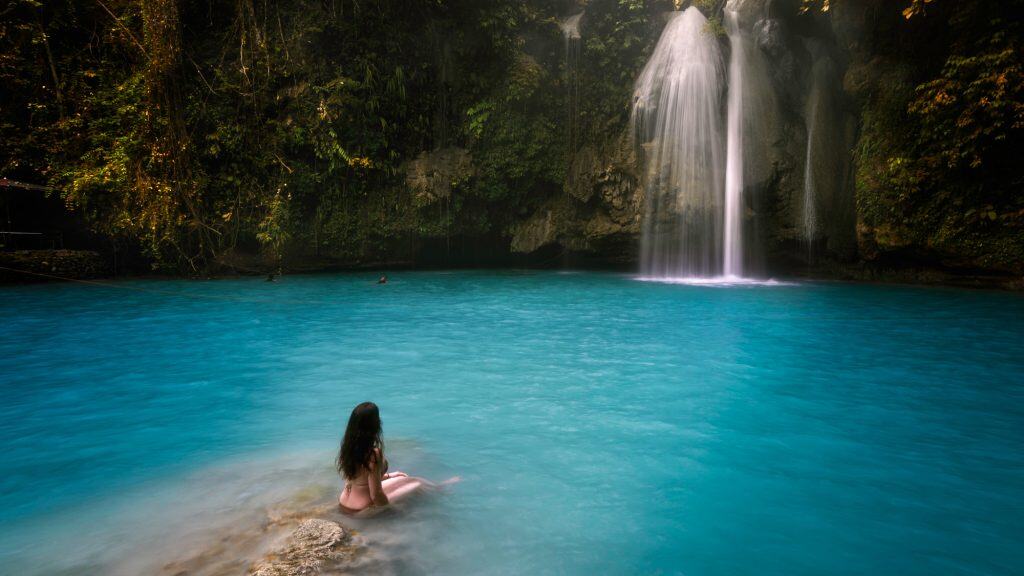 A peaceful scene of a woman sitting on the edge of a turquoise pool, gazing at Kawasan Falls as it spills gently from the rocky cliff surrounded by jungle. The water looks milky and serene under soft light.