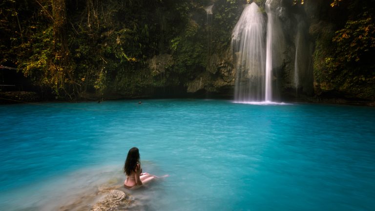 A peaceful scene of a woman sitting on the edge of a turquoise pool, gazing at Kawasan Falls as it spills gently from the rocky cliff surrounded by jungle. The water looks milky and serene under soft light.