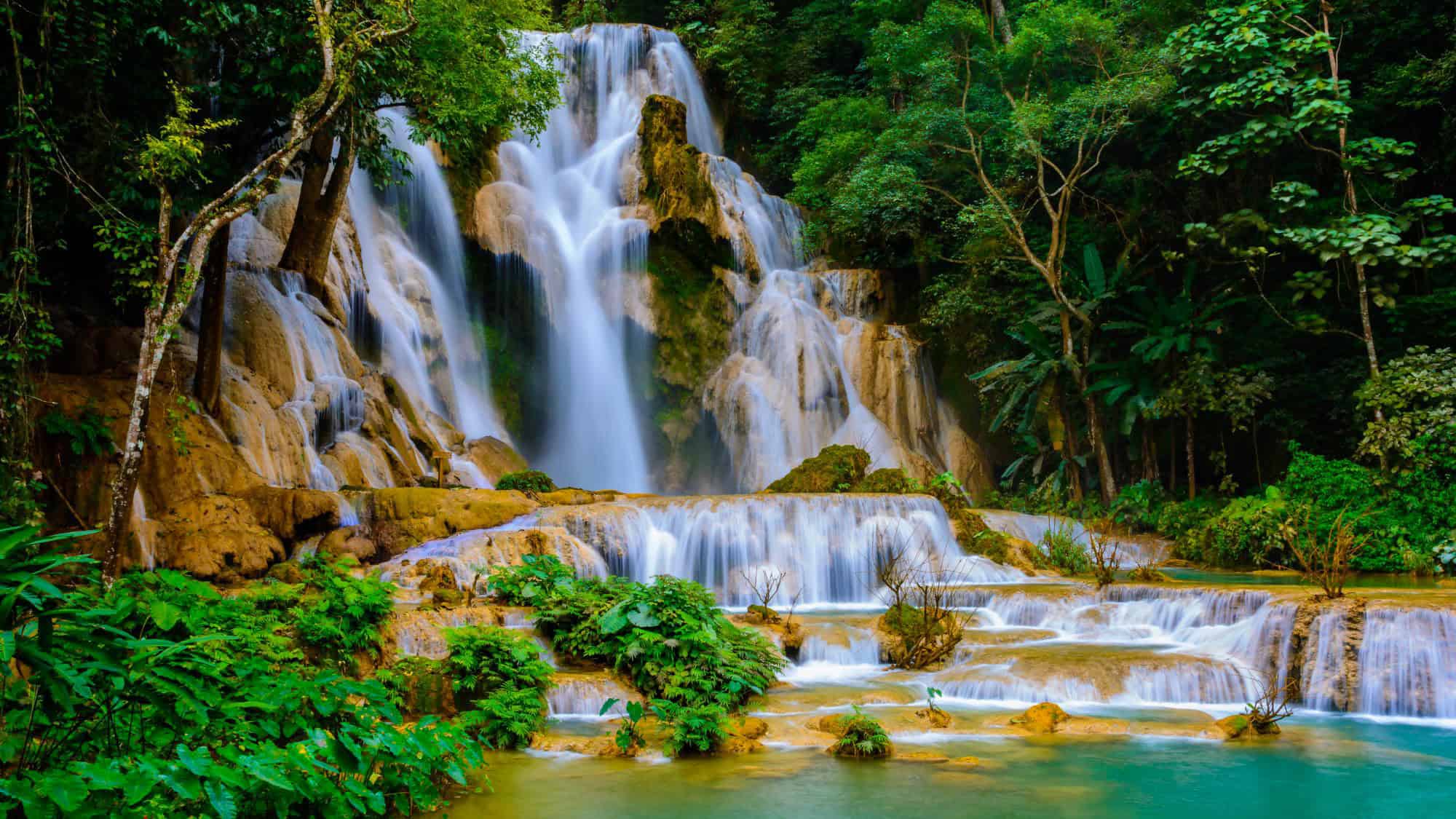 A multi-tiered waterfall tumbles over limestone rocks into a series of emerald-blue pools in the jungles of Laos. The lush foliage frames the scene, adding to the waterfall’s serene, jungle oasis vibe.