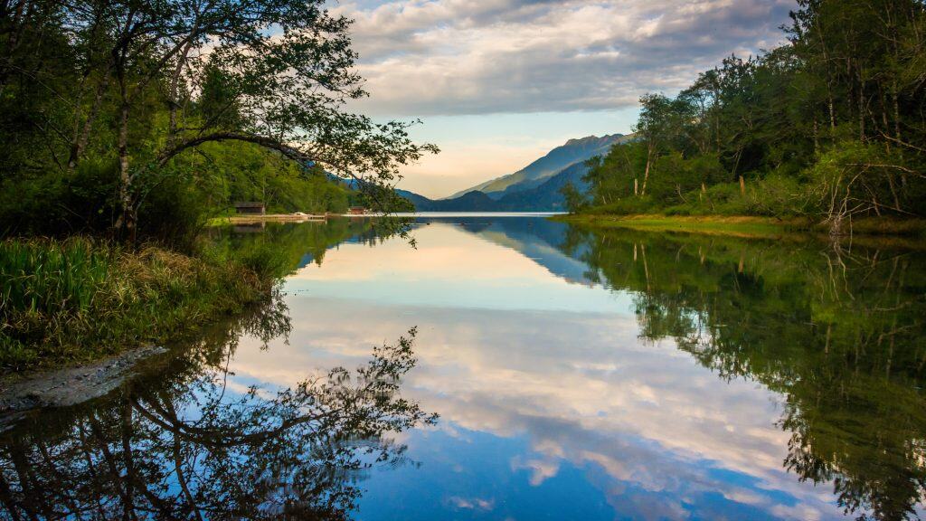 Still waters of Lake Crescent perfectly mirror the surrounding trees, clouds, and distant mountains in the golden light of early morning or late afternoon.