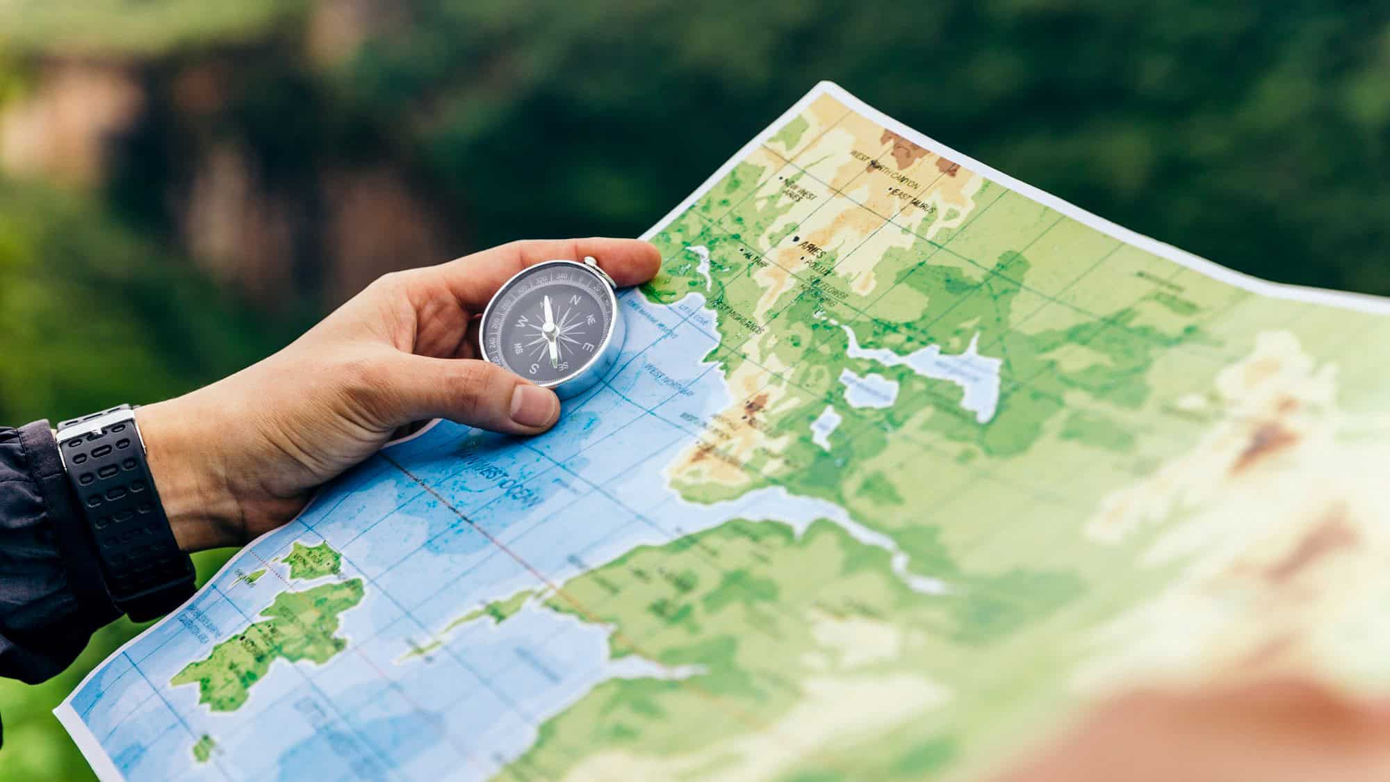 A person’s hand holds a compass above a topographic map, with green landscapes and blue water details, emphasizing outdoor navigation and adventure.