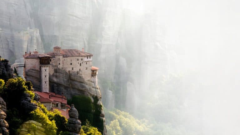A historic stone monastery clings to a towering rock pillar, bathed in soft morning mist, surrounded by the dramatic vertical cliffs of Meteora, Greece.