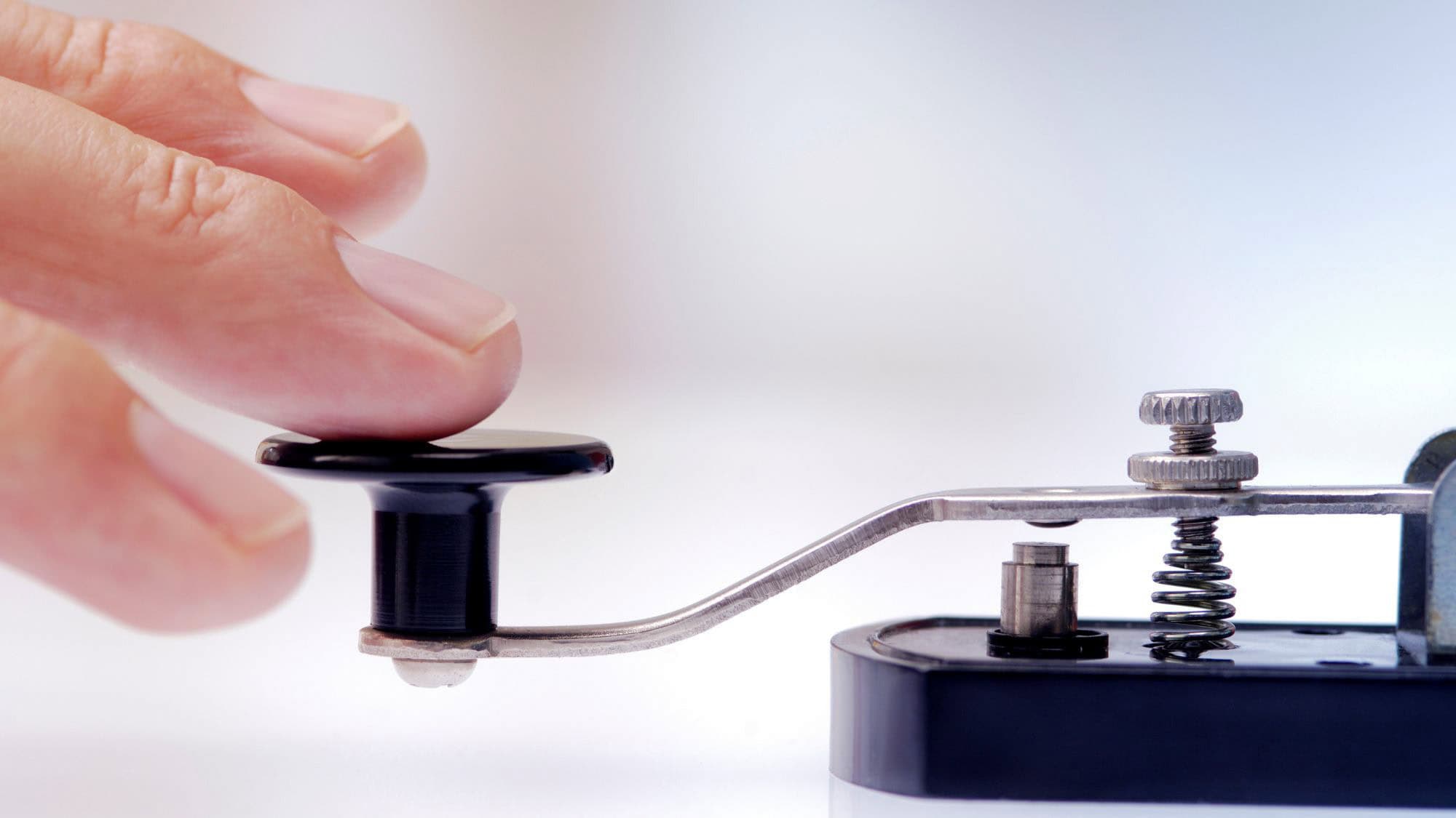 A close-up of a hand pressing down on a vintage black and silver Morse code key, with clear focus on the mechanical spring and button components.