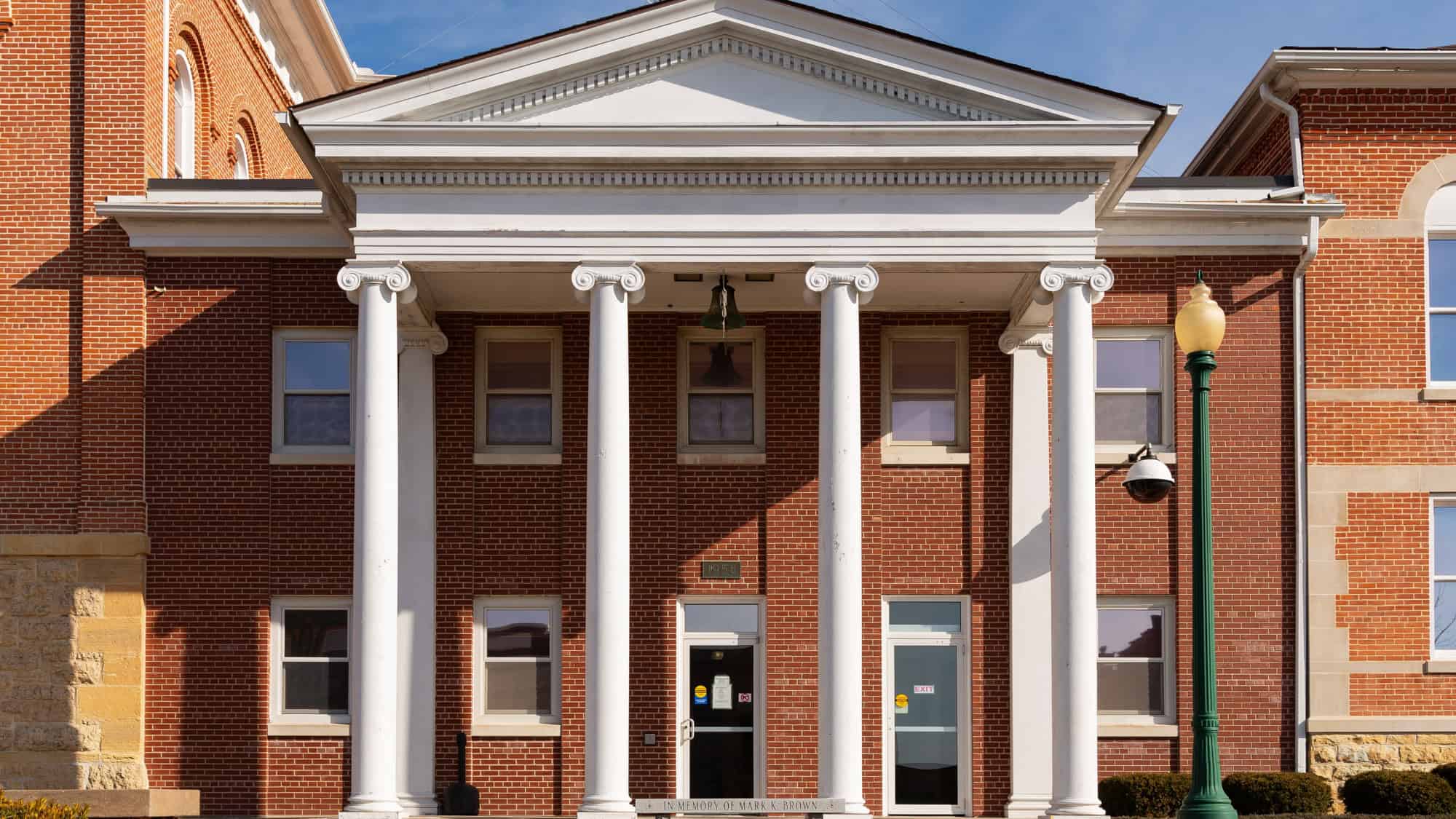 A symmetrical red-brick government building with white columns and a bell over the entryway is pictured under a sunny sky, marked with the year "1958."