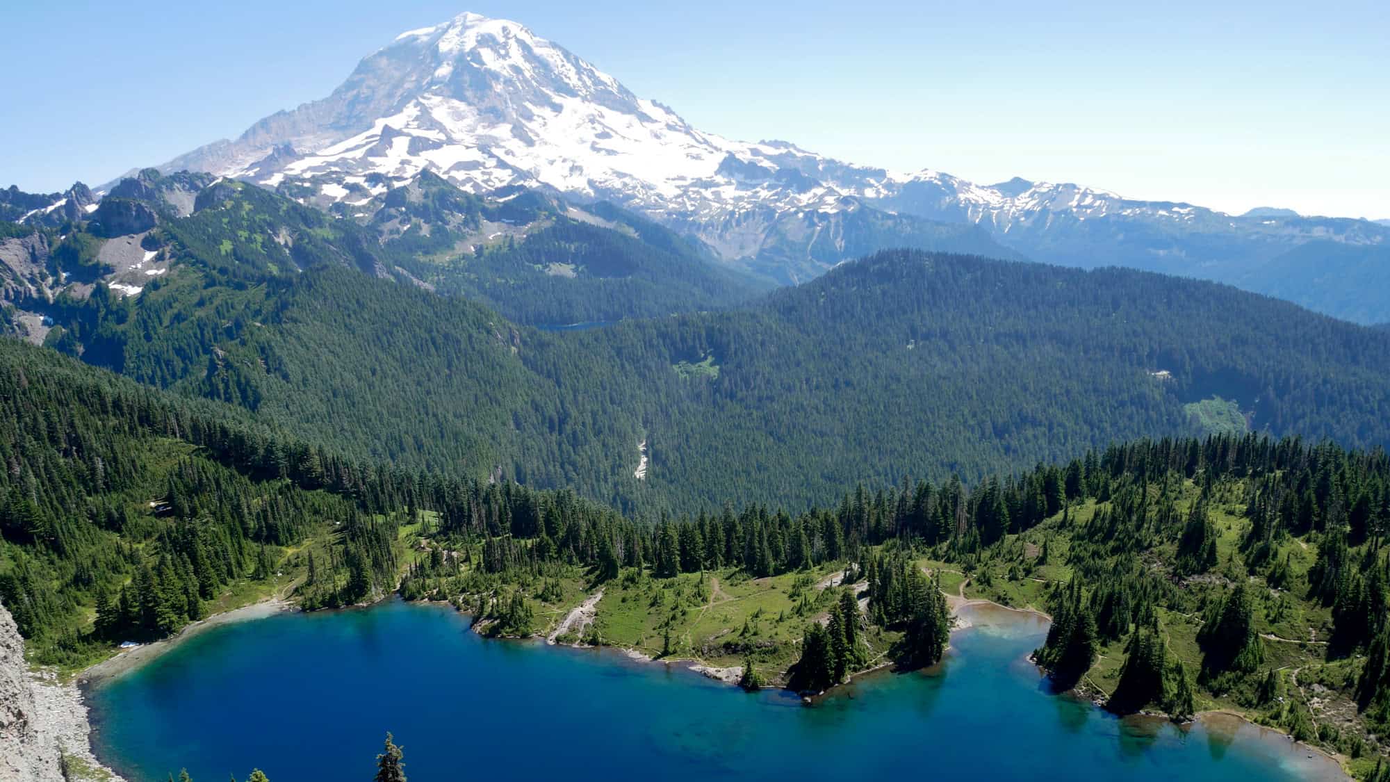 A stunning aerial view of the deep blue Mowich Lake surrounded by thick evergreen forests, with the snow-capped peak of Mount Rainier towering in the distance under a clear blue sky.
