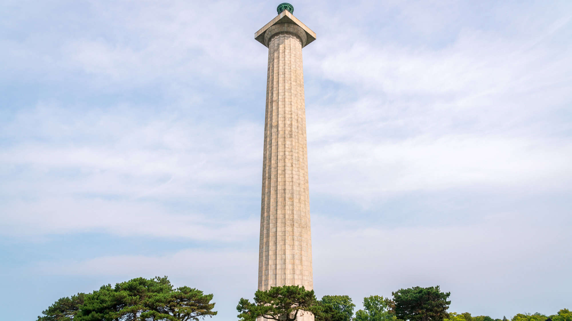 A tall, fluted column made of stone rises high above a line of trees under a partly cloudy sky. This is the Perry’s Victory and International Peace Memorial in Ohio, commemorating the Battle of Lake Erie.